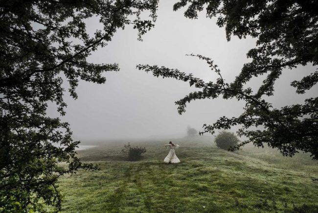 Tanzende Frau in langem, hellem Kleid auf einer Wiese im Nebel.