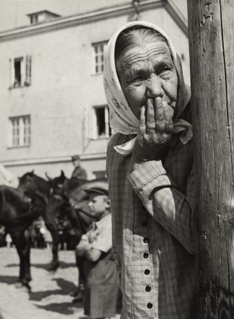 Portrait: alte Frau mit Kopftuch lehnt an einem Holzmast und hält sich die Hand vor den Mund