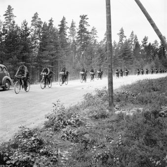 Soldaten fahren auf Fahrrädern eine Landstraße vor einem Wald entlang.