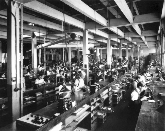 Arbeiter*innen an Arbeitstischen in einer Fabrikhalle.